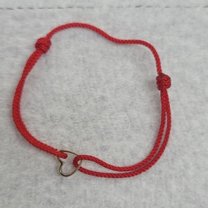 May include: A red cord bracelet with a gold-colored heart charm. The bracelet features adjustable knots for sizing. The heart charm is a simple, elegant design. The bracelet is displayed on a light gray surface.