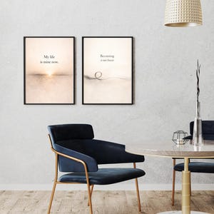 May include: Two framed art prints with inspirational quotes hang on a wall. One says "My life is mine now." The other says "Becoming is not linear." A blue chair with gold legs and a round table are in the foreground.