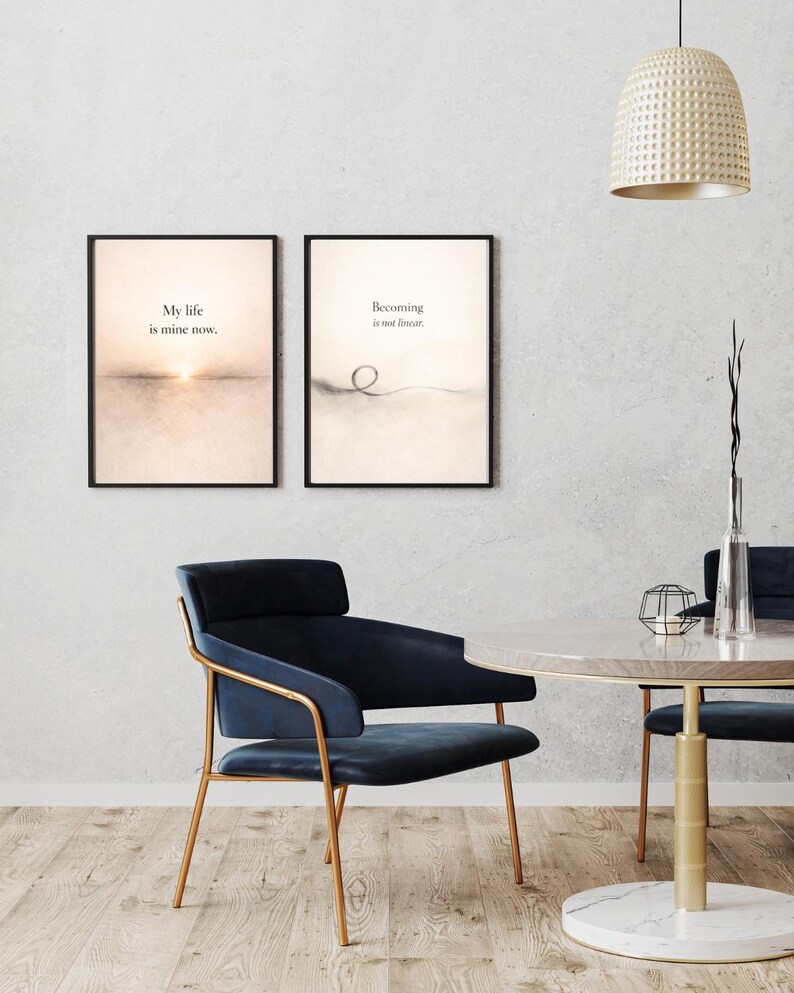 May include: Two framed art prints with inspirational quotes hang on a wall. One says "My life is mine now." The other says "Becoming is not linear." A blue chair with gold legs and a round table are in the foreground.
