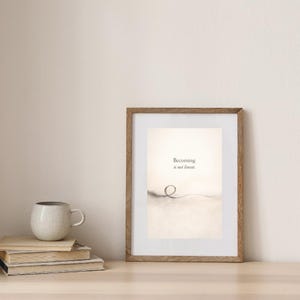 May include: A framed print with the text "Becoming is not linear." The artwork features a minimalist design with a looping line. A stack of books and a speckled mug are on a wooden surface.
