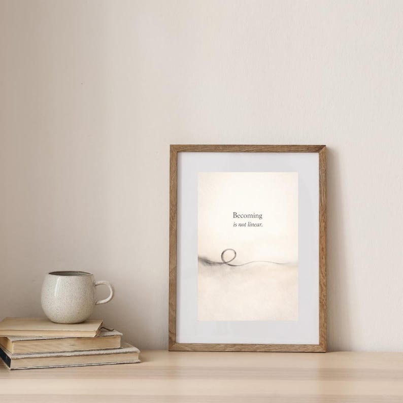May include: A framed print with the text "Becoming is not linear." The artwork features a minimalist design with a looping line. A stack of books and a speckled mug are on a wooden surface.