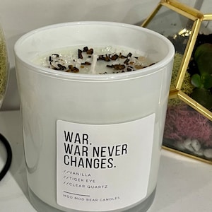 May include: A white glass candle with the text "WAR. WAR NEVER CHANGES." and the scent "Vanilla". The candle is topped with clear quartz and tiger eye stones. The brand name "MOO MOO BEAR CANDLES" is printed on the label.