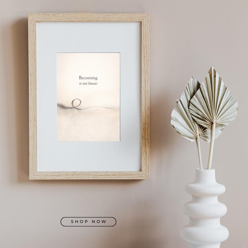 May include: A framed print with the text "Becoming is not linear." The artwork features a minimalist design with a light background and a simple line drawing. A white vase with dried palm leaves is to the right.