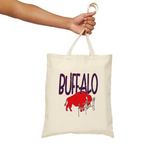 Peut inclure: Sac fourre-tout crème avec le mot "BUFFALO" en bleu, lettrage vieilli. Un bison rouge est en dessous, avec le mot "football" en écriture cursive. Le sac est tenu par une main.
