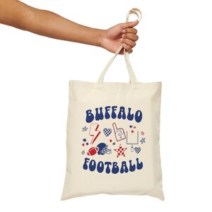 Peut inclure: Sac fourre-tout crème avec les mots "BUFFALO FOOTBALL" en bleu, avec des graphiques rouges et bleus d'un ballon de football, d'un casque, d'un éclair et d'étoiles. Le sac a deux poignées et est tenu par une main.