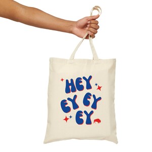 Peut inclure: Sac fourre-tout crème avec le texte "HEY EY EY EY" en lettres bleues à bulles, agrémenté d'étoiles rouges et d'un logo de bison rouge. Le sac est tenu par une main sur fond blanc.