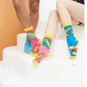 May include: A pair of socks with a comic book theme. One sock is pink with yellow and blue accents and says "OMG" in a speech bubble. The other sock is blue with yellow and white accents and says "POW" in a speech bubble.