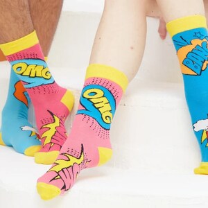 May include: A pair of colorful socks with a comic book theme. One sock features the word "OMG" in a blue speech bubble with yellow and red accents. The other sock features the word "POW" in a red speech bubble with yellow and white accents.
