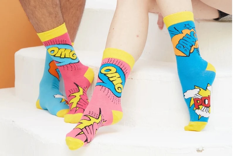 May include: A pair of colorful socks with a comic book theme. One sock features the word "OMG" in a blue speech bubble with yellow and red accents. The other sock features the word "POW" in a red speech bubble with yellow and white accents.
