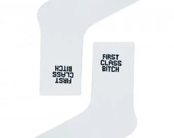 May include: Two white socks with the text "FIRST CLASS BITCH" in black. The word "CARNAVAL" is printed in black on the top of one sock. The socks are a casual style.