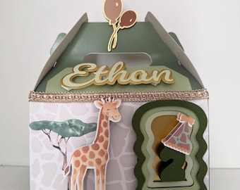 May include: A decorative party favor box with a safari theme. The box is green and white with a giraffe and tree design. The name "Ethan" is displayed on the front, along with a balloon decoration. The box has a handle and a decorative trim.