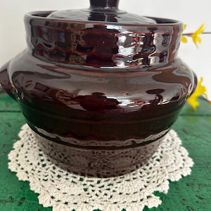 Pode incluir: Uma panela de cerâmica marrom escura com tampa e acabamento brilhante. A panela tem formato arredondado com duas pequenas alças e fica sobre uma toalha de crochê creme. A tampa tem um pequeno botão na parte superior.