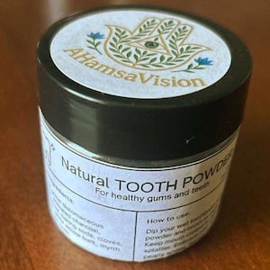 May include: A small, black-lidded jar of natural tooth powder. The label reads "Natural TOOTH POWDER" with the text "For healthy gums and teeth." The jar's lid features a white label with the text "AHamsaVision" and a hand symbol.