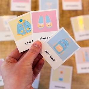 Op de afbeelding: Een hand die drie geïllustreerde flashcards vasthoudt. De kaarten tonen een blauwe jas, blauwe schoenen en een blauwe rugzak. Elke kaart heeft een woordlabel: "coat", "shoes" en "pack back". De achtergrond bevat andere kaarten.
