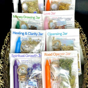 May include: Assortment of packaged jars with labels like "Protection Jar," "Confidence Jar," and "Love Jar." Each jar contains various items such as candles, herbs, and crystals. The jars are arranged on a decorative, gold-colored tray.