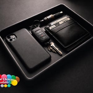 May include: A rectangular black valet tray containing a black smartphone in a case, a black car key fob with metal keys, a black leather wallet with visible cards, and a small black and silver torch. The text Color Choices Available appears.