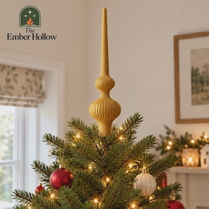 May include: A mustard yellow, ribbed, finial-style Christmas tree topper with a tall, pointed spire and a bulbous base. The topper is positioned at the very top of a green pine tree decorated with warm white string lights and red ornaments.