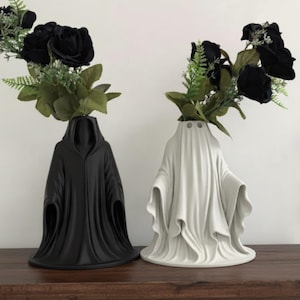 May include: Two decorative vases, one black and one white, each shaped like a draped figure. The black vase resembles a hooded cloak, while the white vase is styled like a ghost. Both hold black roses and green foliage, creating a striking contrast.