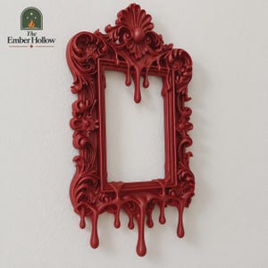 May include: A deep red rectangular picture frame featuring ornate, baroque-style scrollwork and decorative carvings. The frame design includes a distinctive dripping effect along the bottom and top edges, creating a fluid, melting appearance.