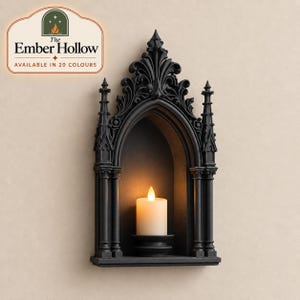 May include: A black wall-mounted candle holder featuring an arched frame with intricate architectural detailing and pointed spires. A single white pillar candle with a warm glowing flame sits on the base inside the arched alcove.