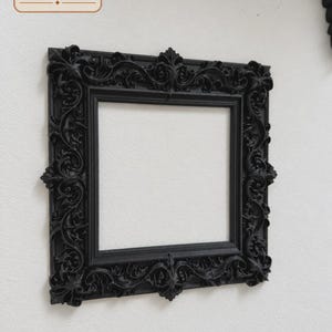 May include: A square black picture frame featuring an ornate, raised relief pattern of scrolling leaves and floral motifs along the border. The frame has a matte finish and a hollow centre. The logo for The Ember Hollow is visible in the corner.
