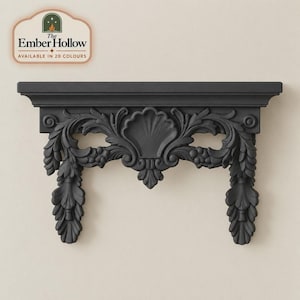 May include: A matte black wall shelf featuring an ornate carved design with a central shell motif, scrolling acanthus leaves, and two hanging floral pendants. The top surface is flat and rectangular. A logo reads The Ember Hollow AVAILABLE IN 20 COLOURS.