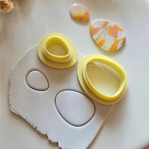 May include: A craft scene featuring polymer clay and tools. There are yellow oval cutters, white clay with oval cutouts, and two finished clay pieces with orange and white marbling. Dried flowers are in the background.