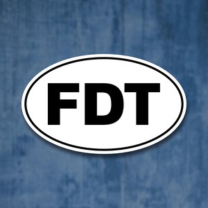 Op de afbeelding: Witte ovale sticker met de tekst "FDT" in zwarte letters.