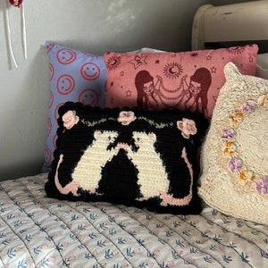 May include: A bed with several decorative pillows. One pillow is black with white cat figures and pink flowers. Other pillows are pink and blue with various designs. The bedspread has a floral pattern.