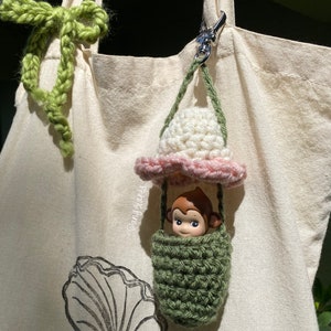 K&ouml;nnte beinhalten: Ein geh&auml;kelter gr&uuml;ner und rosa Hei&szlig;luftballon-Anh&auml;nger mit einer kleinen Affenfigur im Inneren. Der Anh&auml;nger ist an einer wei&szlig;en Tragetasche mit einer gr&uuml;nen geh&auml;kelten Schleife befestigt.