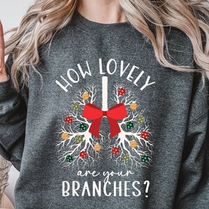 Puede incluir: Sudadera gris oscuro con el texto "HOW LOVELY are your BRANCHES?" en blanco. El diseño presenta un gráfico estilizado de pulmones con un lazo rojo y adornos coloridos. La sudadera es de estilo cómodo e informal.