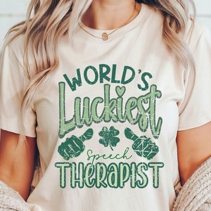 May include: A beige t-shirt with the text "WORLD'S Luckiest Speech Therapist" in green. The design includes a shamrock and two green hand gestures. The shirt is made of a soft material.