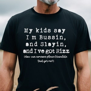 May include: Black t-shirt with white text that reads "My kids say I'm Bussin, and Slayin, and I've got Rizz (Now can someone please translate that for me?)". The shirt is worn by a person standing in a field.