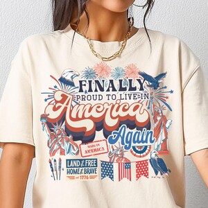 Puede incluir: Una camiseta beige con un estampado gráfico de fuegos artificiales, estrellas y el texto "FINALLY PROUD TO LIVE IN AMERICA Again". El gráfico también incluye el texto "MADE IN AMERICA", "LAND of FREE HOME of BRAVE", y "1776".