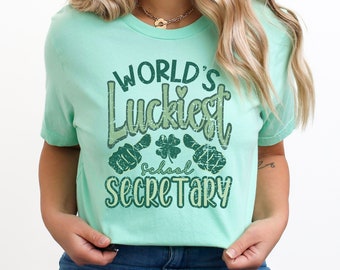 May include: A light green t-shirt with the text "World's Luckiest School Secretary" in a green and white distressed font. The text is surrounded by green shamrocks and thumbs up.