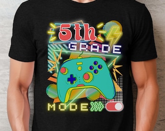 Puede incluir: Camiseta negra con un diseño de neón que presenta el texto "5th GRADE MODE" y un mando de videojuegos turquesa. El diseño incluye rayos amarillos y un interruptor basculante rojo y blanco.