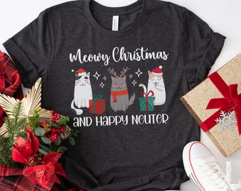 Könnte beinhalten: Dunkelgraues T-Shirt mit dem Text "Meowy Christmas and Happy Neuter". Das Shirt zeigt eine Grafik von drei Katzen mit Weihnachtsmützen und Schals, mit Geschenkverpackungen. Weiße Turnschuhe und eine Geschenkbox sind ebenfalls zu sehen.