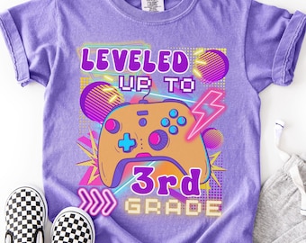 May include: A purple Comfort Colors t-shirt with a video game controller graphic. The shirt features the text "LEVELED UP TO 3rd GRADE" in a neon-style design. Black and white checkered slip-on shoes are visible.