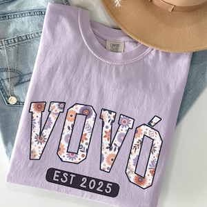 Puede incluir: Una camiseta lavanda con un diseño de estampado floral que dice "VOVO EST 2025".