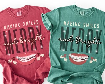 May include: Two t-shirts, one red and one teal, with the text "MAKING SMILES MERRY and BRIGHT". Each shirt features a graphic of a mouth with braces and holly leaves. The shirts are casual and festive.