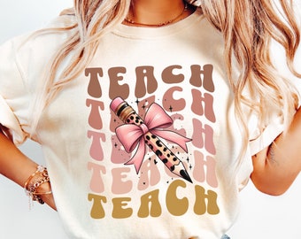 May include: A white t-shirt with the word "TEACH" repeated in a retro font in brown, pink, and gold colors. A pencil with a pink bow and leopard print is centered on the shirt.