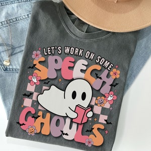 May include: Dark gray t-shirt with the text "LET'S WORK ON SOME SPEECH GHOULS" in colorful bubble letters. A cartoon ghost reading a book is in the center. The shirt is styled with a tan hat and blue jeans.