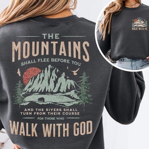 May include: Dark gray sweatshirt with a mountain graphic and the text "The Mountains Shall Flee Before You" and "Walk With God." The design includes a red sun, bear, and trees. A smaller graphic with "Walk With Me" is on the upper right.