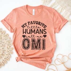 May include: A peach-colored t-shirt with the text "My favorite little humans call me OMI" printed in black. The text has a distressed, vintage look.