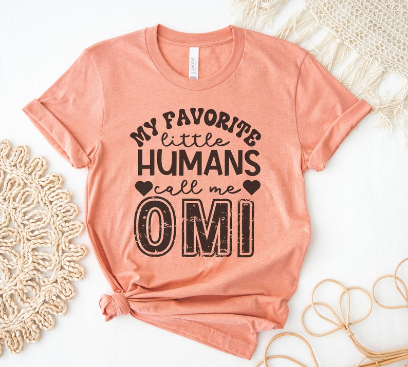 May include: A peach-colored t-shirt with the text "My favorite little humans call me OMI" printed in black. The text has a distressed, vintage look.