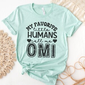 May include: A light blue t-shirt with the text "My favorite little humans call me Omi" in black with a distressed font. The text is centered on the shirt and includes two small black hearts.