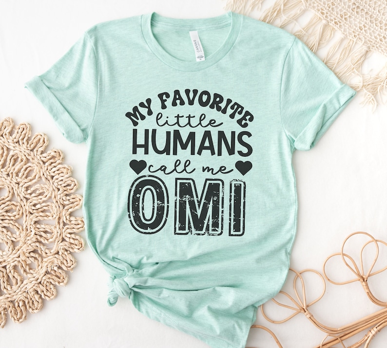 May include: A light blue t-shirt with the text "My favorite little humans call me Omi" in black with a distressed font. The text is centered on the shirt and includes two small black hearts.