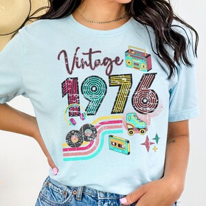 May include: Light blue t-shirt with a retro design featuring the word "Vintage" above the number "1976". The design includes a boombox, roller skate, cassette tape, and records. The shirt has a crew neck.
