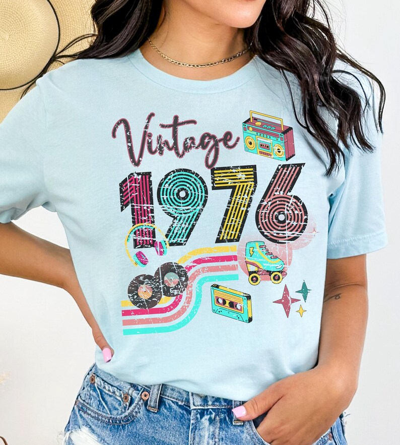 May include: Light blue t-shirt with a retro design featuring the word "Vintage" above the number "1976". The design includes a boombox, roller skate, cassette tape, and records. The shirt has a crew neck.