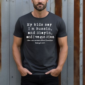 May include: Dark gray t-shirt with white text that reads "My kids say I'm Bussin, and Slayin, and I've got Rizz (Now can someone please translate that for me?)". The shirt is a crew neck style.